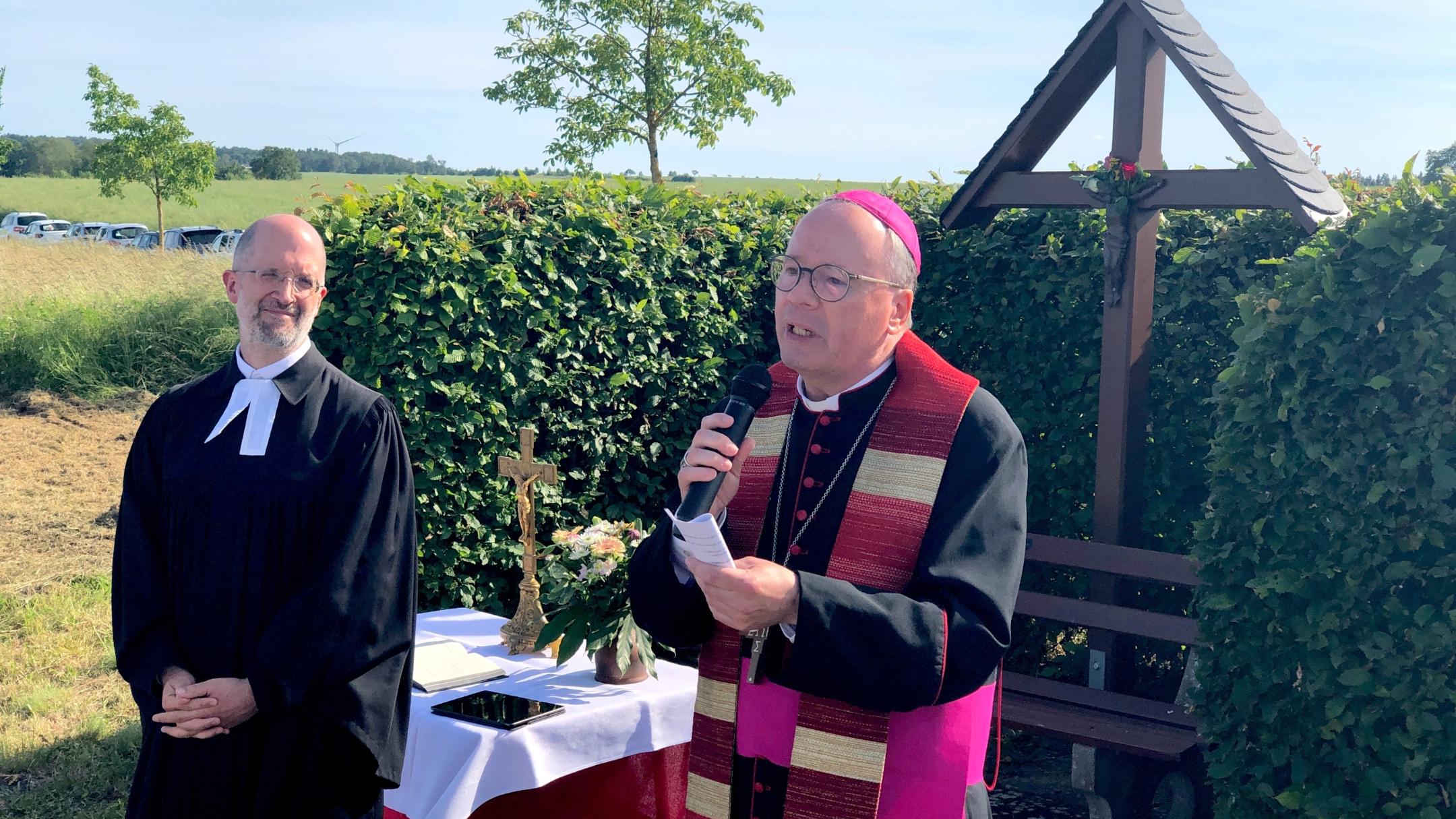 evangelischer und katholischer Geistlicher im Gespräch vor einem Kreuz und einer Hecke