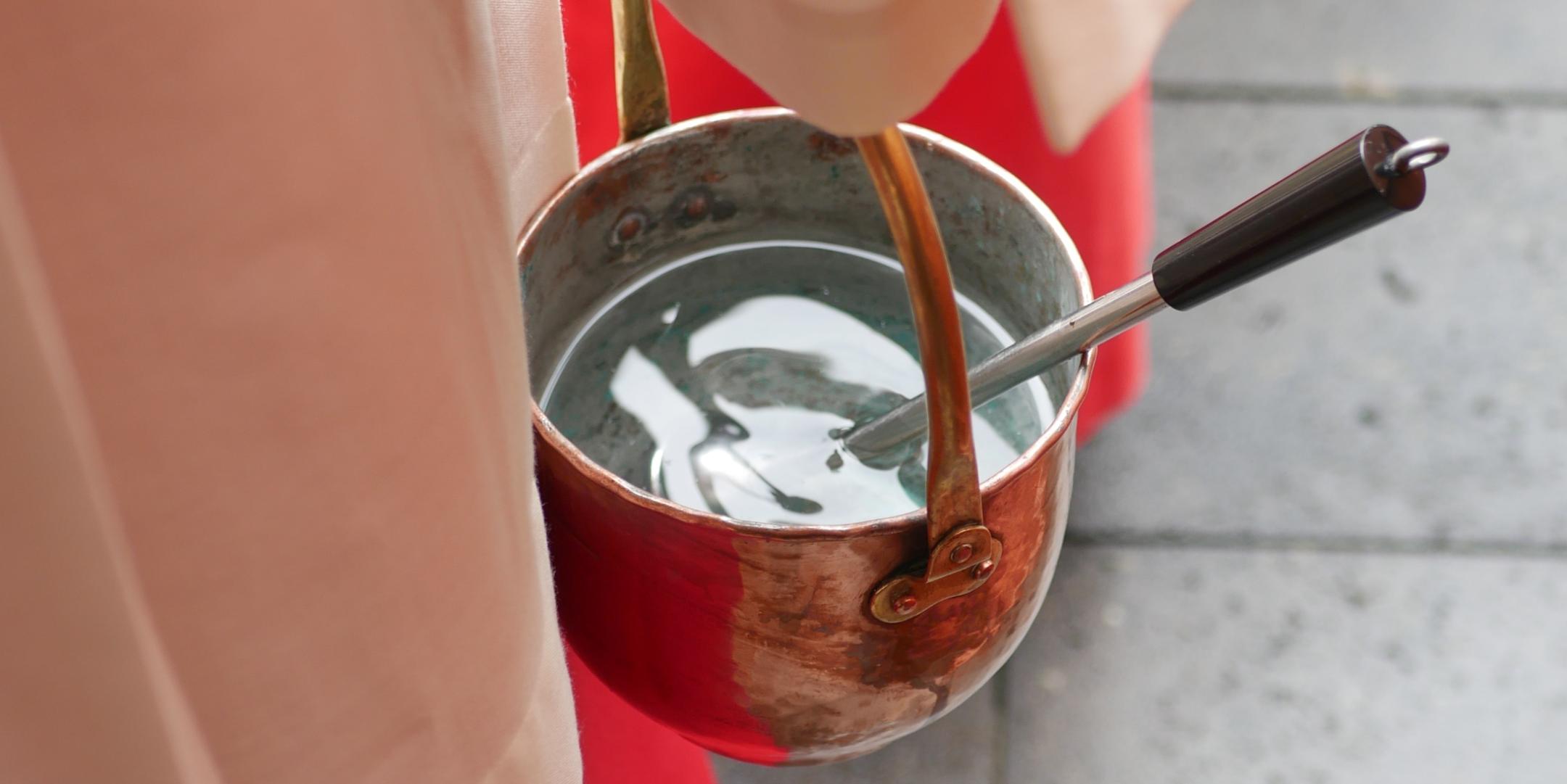 Eine Person trägt ein rotes liturgisches Gewand. In der Hand hält die Person ein kleines Metallgefäß mit Weihwasser. Darin steckt ein Weihwassersprenger mit schwarzem Griff. Das Gefäß hängt an einem kurzen Henkel. Der Boden aus grauen Steinplatten ist im Hintergrund zu sehen. Die Aufnahme zeigt einen engen Ausschnitt und konzentriert sich auf das Gefäß und das Gewand. (c) Lars F. Reiplinger Eine Person trägt ein rotes liturgisches Gewand. In der Hand hält die Person ein kleines Metallgefäß mit Weihwasser. Darin steckt ein Weihwassersprenger mit schwarzem Griff. Das Gefäß hängt an einem kurzen Henkel. Der Boden aus grauen Steinplatten ist im Hintergrund zu sehen. Die Aufnahme zeigt einen engen Ausschnitt und konzentriert sich auf das Gefäß und das Gewand.