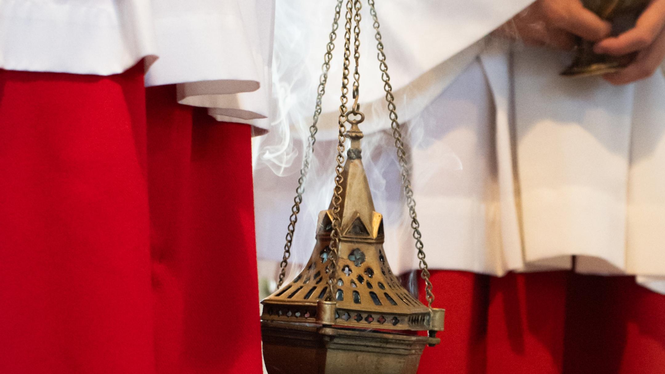 Ein Weihrauchfass aus Metall hängt an Ketten und ist scharf im Vordergrund abgebildet. Im Hintergrund sind unscharf liturgische Gewänder in Weiß und Rot zu erkennen.