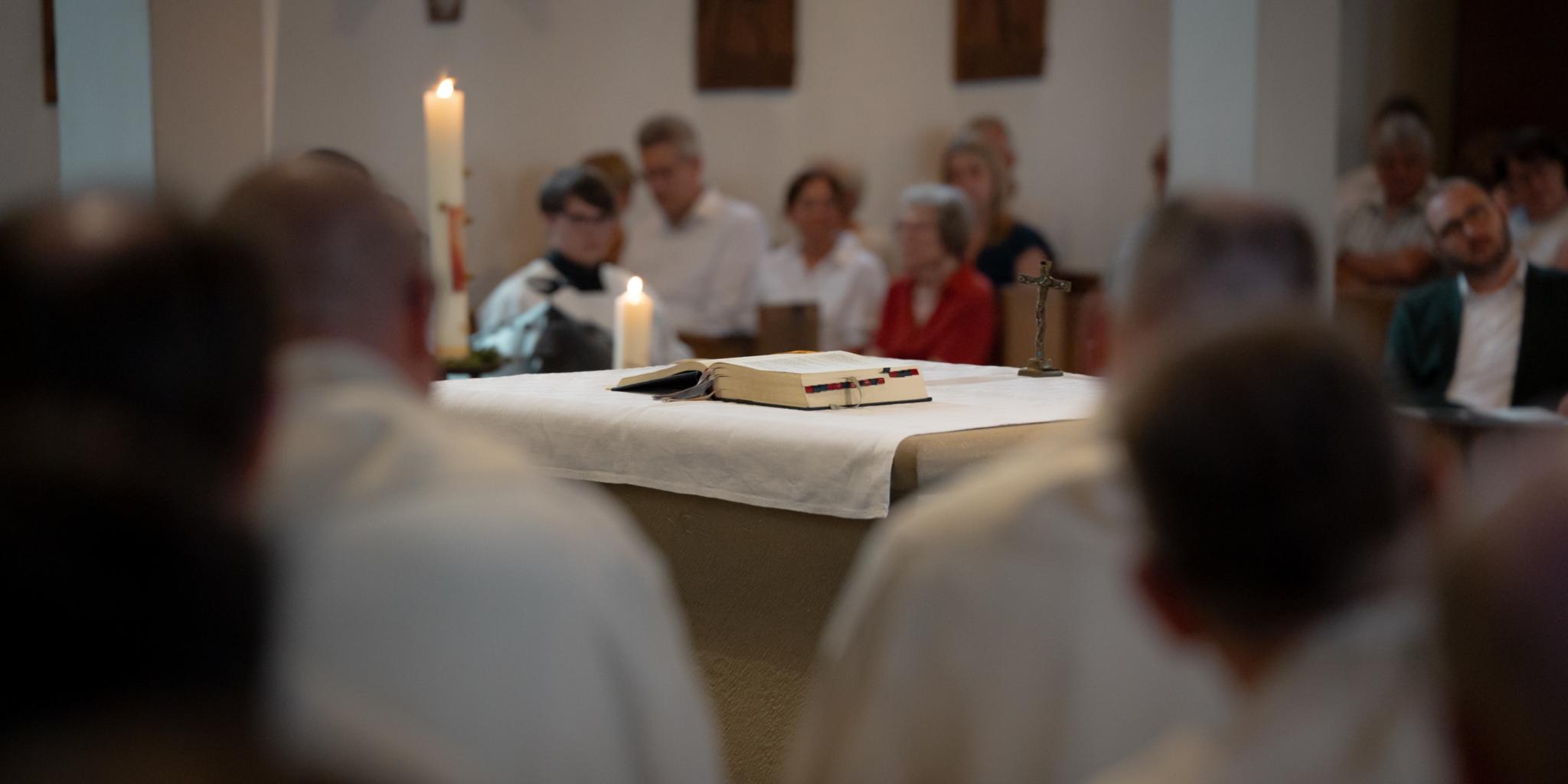 Ein Altar mit weißem Altartuch steht im Mittelpunkt der Aufnahme. Darauf liegen ein geöffnetes Messbuch und ein kleines Kreuz. Im Hintergrund sitzen Besucherinnen und Besucher des Gottesdienstes, während Kerzen den Altarbereich rahmen.