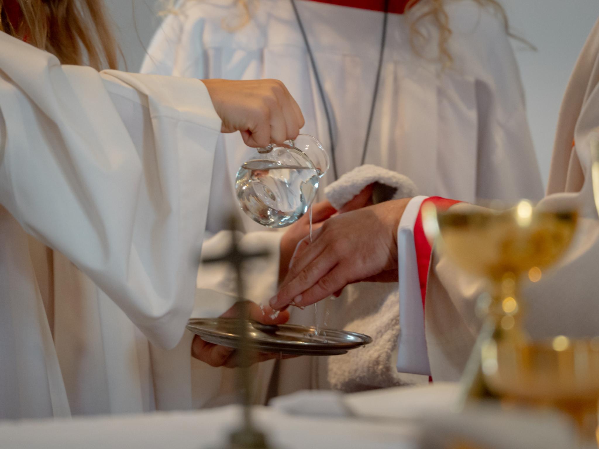 Handwaschung im Gottesdienst Eine Person gießt Wasser aus einem Glaskännchen über die Hände einer anderen Person. Unter den Händen wird eine Schale gehalten. Die liturgische Handwaschung findet am Altar während der Messfeier statt.