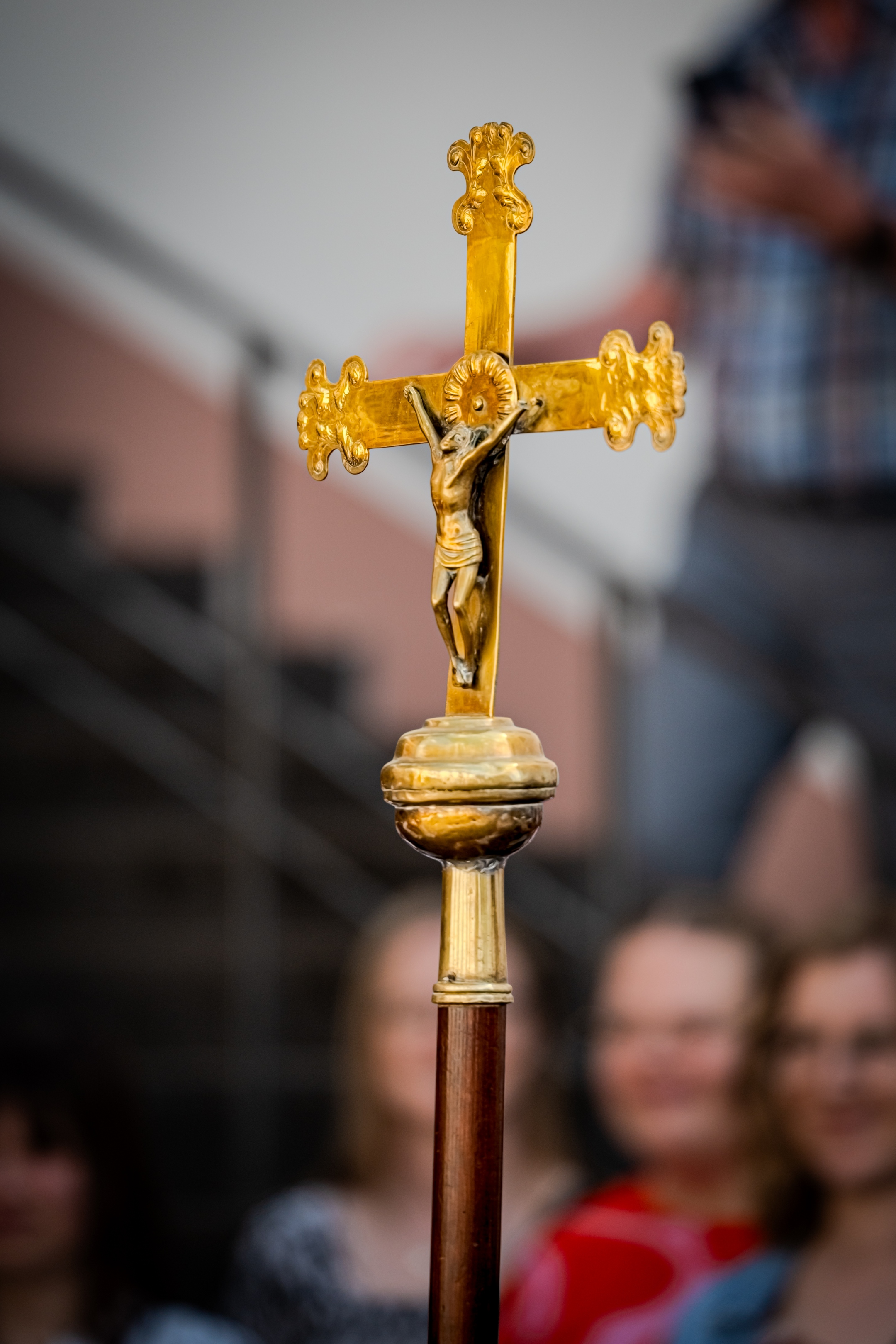 Ein goldenes Prozessionskreuz mit Christusdarstellung steht im Mittelpunkt des Bildes. Im unscharfen Hintergrund sind Besucherinnen und Besucher eines Gottesdienstes zu erkennen.