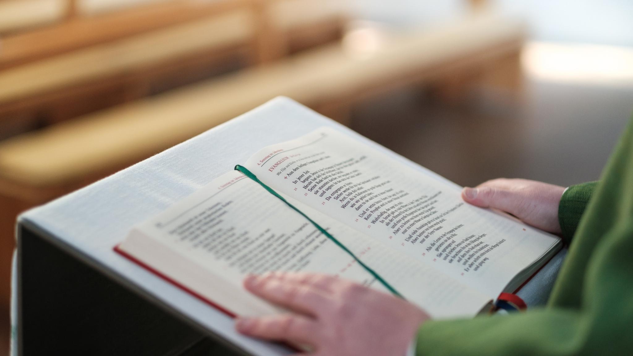Ein aufgeschlagenes Lektionar liegt auf einem Lesepult in einer Kirche. Die Seiten sind gut sichtbar, ein grünes Lesebändchen markiert eine Stelle im Text. Eine Hand hält das Buch offen. Im Hintergrund sind mehrere hölzerne Kirchenbänke zu sehen, die nach hinten hin unscharf wirken. Der Kirchenraum ist in warmes, ruhiges Licht getaucht.