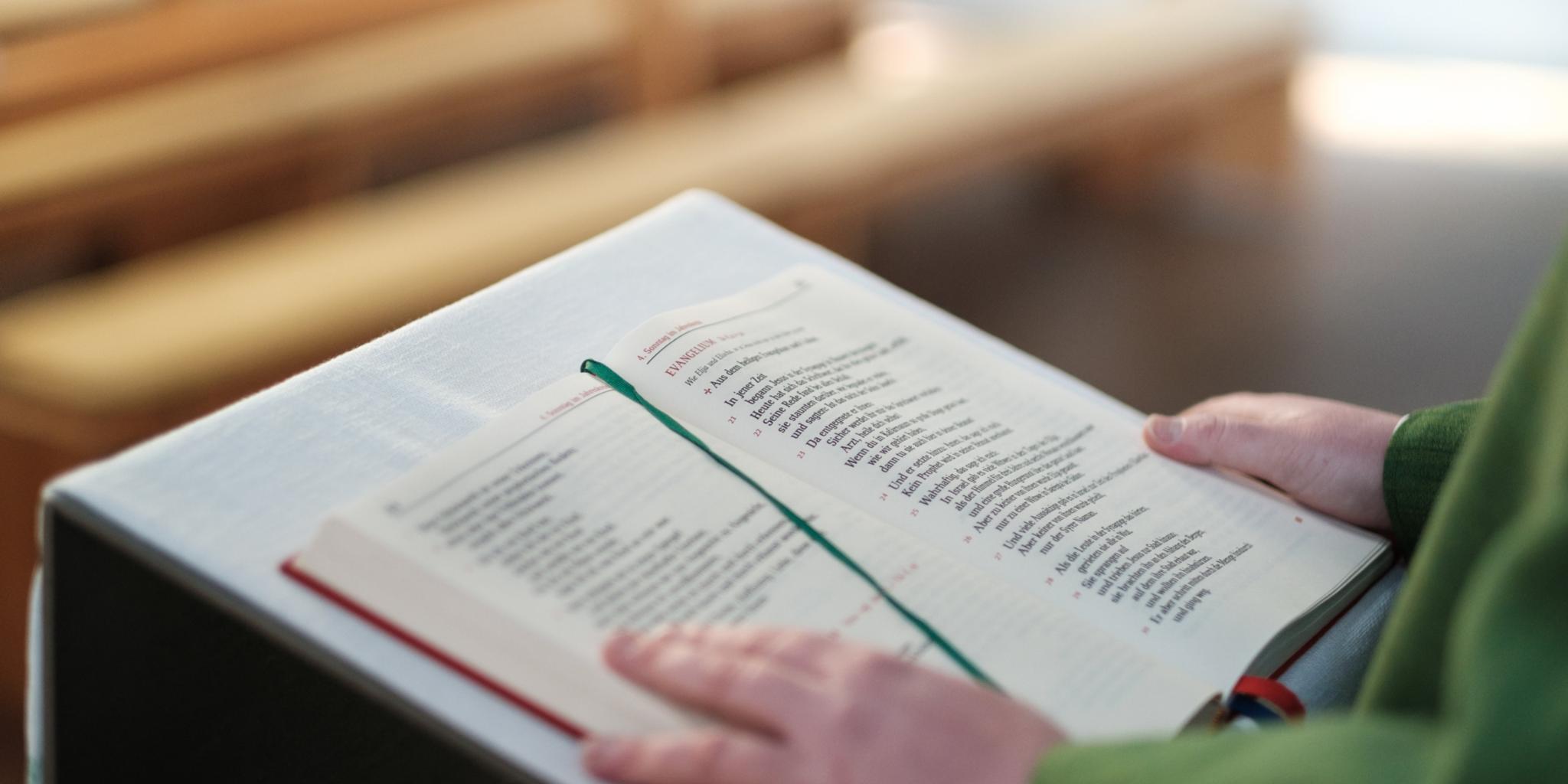 Ein aufgeschlagenes Lektionar liegt auf einem Lesepult in einer Kirche. Die Seiten sind gut sichtbar, ein grünes Lesebändchen markiert eine Stelle im Text. Eine Hand hält das Buch offen. Im Hintergrund sind mehrere hölzerne Kirchenbänke zu sehen, die nach hinten hin unscharf wirken. Der Kirchenraum ist in warmes, ruhiges Licht getaucht.