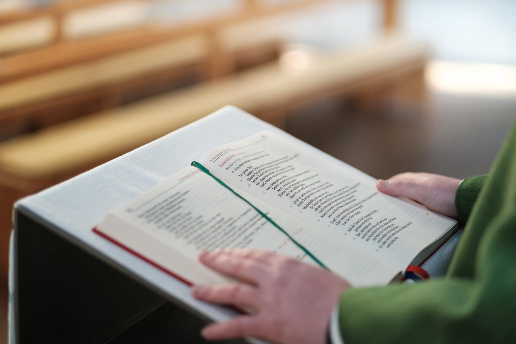 Ein aufgeschlagenes Lektionar liegt auf einem Lesepult in einer Kirche. Die Seiten sind gut sichtbar, ein grünes Lesebändchen markiert eine Stelle im Text. Eine Hand hält das Buch offen. Im Hintergrund sind mehrere hölzerne Kirchenbänke zu sehen, die nach hinten hin unscharf wirken. Der Kirchenraum ist in warmes, ruhiges Licht getaucht.