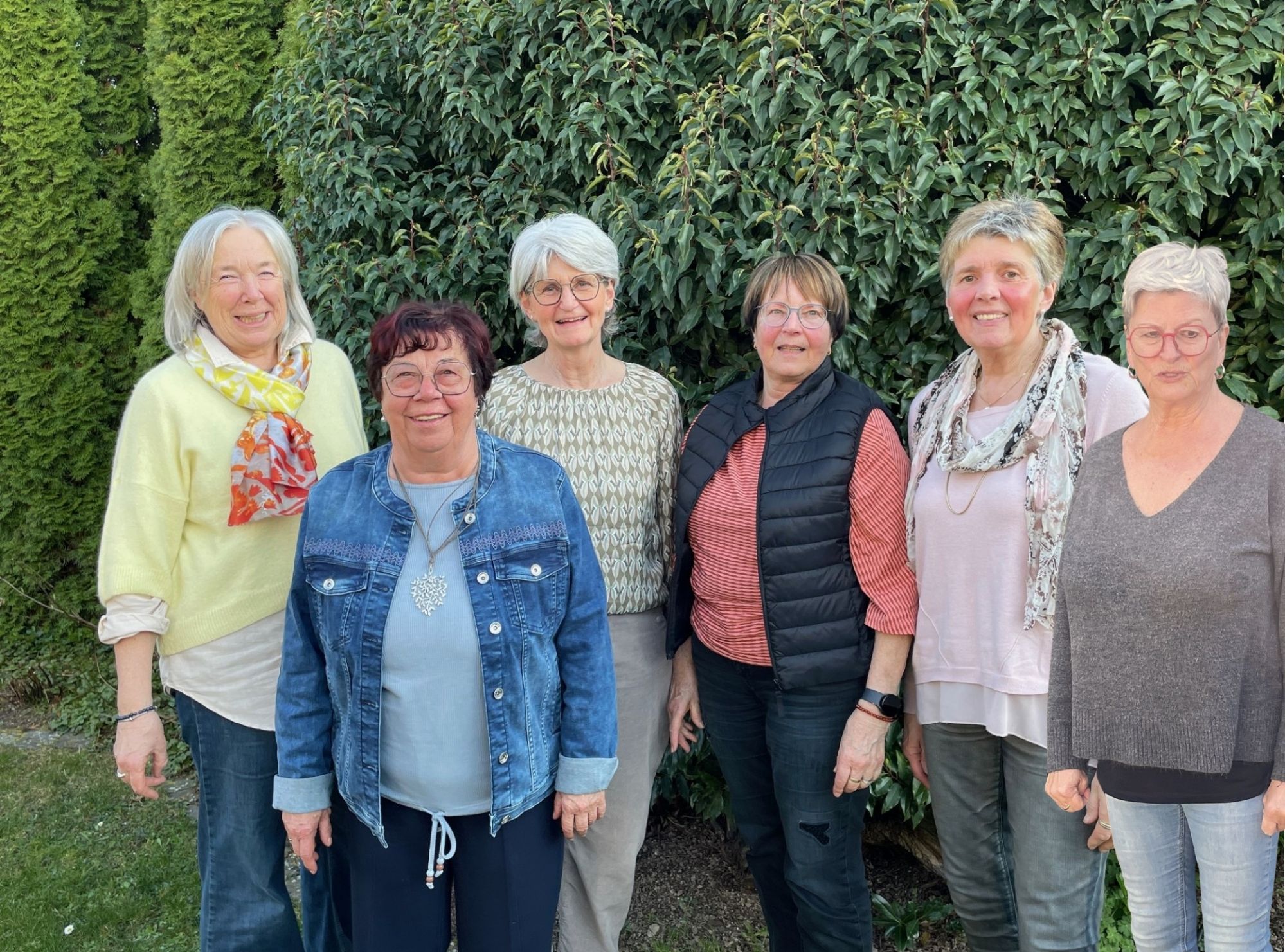 Sechs Frauen stehen lächelnd vor einer Hecke