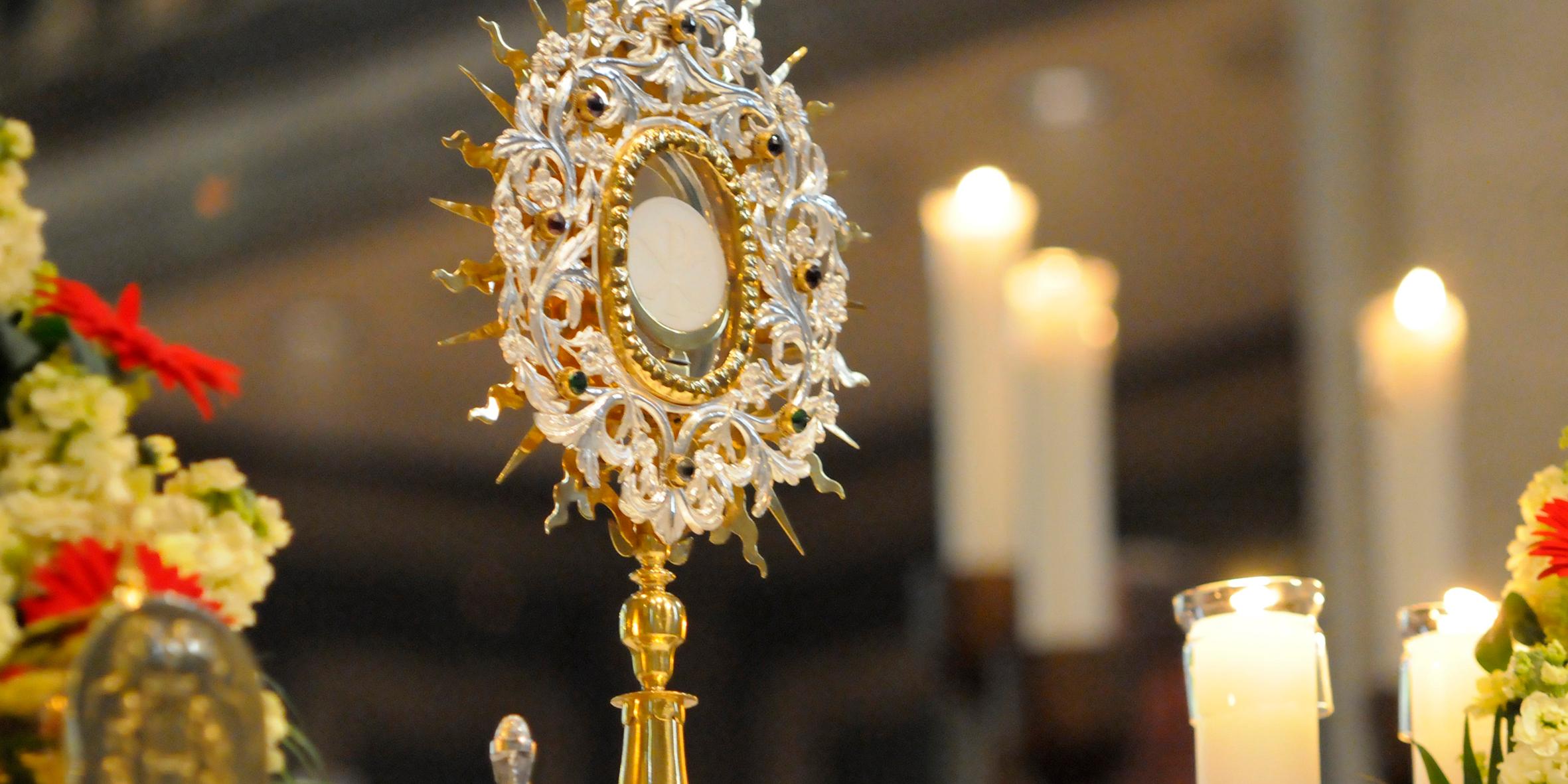 Eine reich verzierte Monstranz steht auf einem Altar in einer Kirche. In der Mitte der Monstranz ist die weiße Hostie sichtbar. Die Monstranz besteht aus gold- und silberfarbenem Metall mit strahlenförmigen Ornamenten. Rundherum stehen mehrere brennende Kerzen in Glasgefäßen. Blumenschmuck rahmt den Altar. Der Hintergrund ist unscharf und zeigt das Innere des Kirchenraums.
