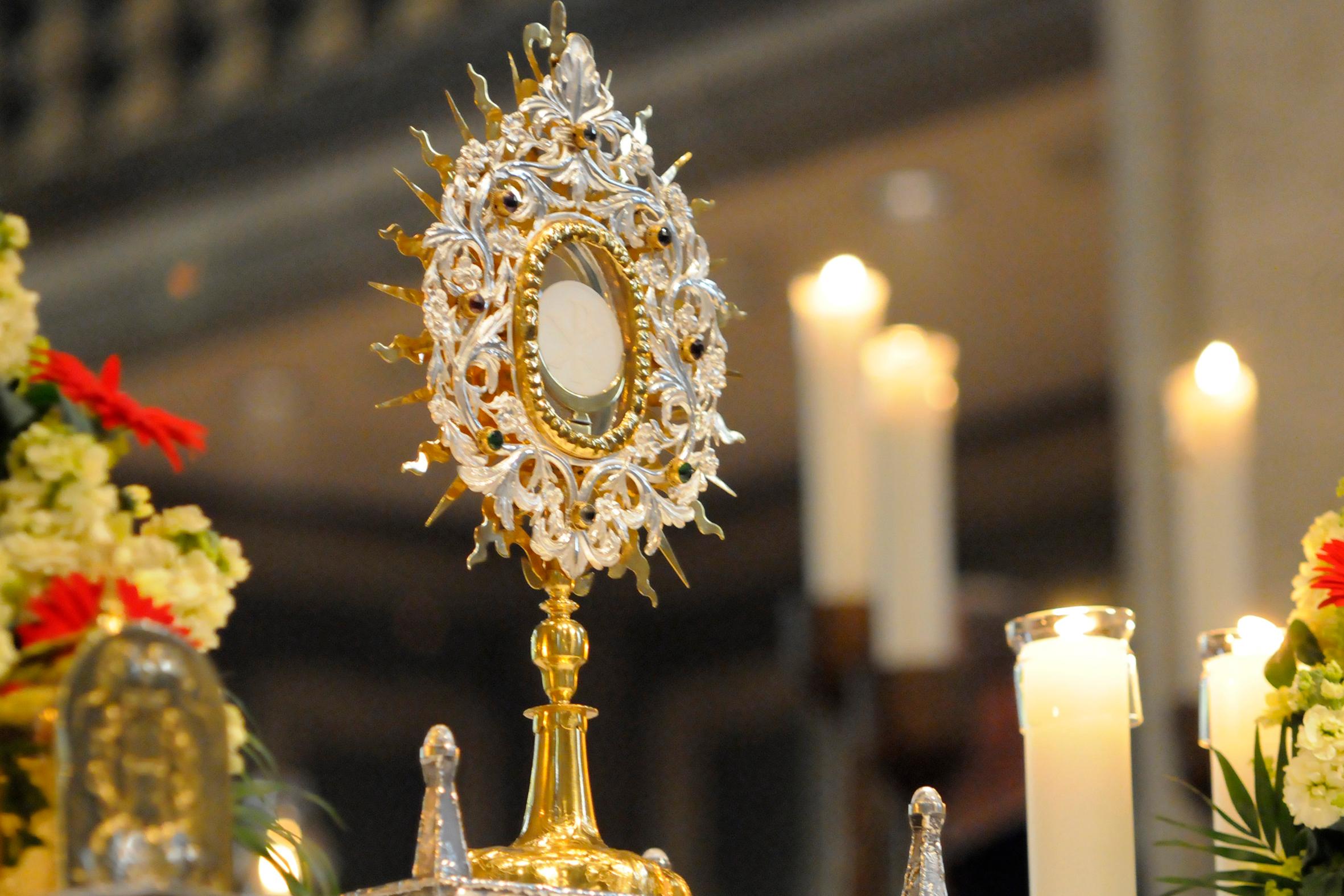 Eine reich verzierte Monstranz steht auf einem Altar in einer Kirche. In der Mitte der Monstranz ist die weiße Hostie sichtbar. Die Monstranz besteht aus gold- und silberfarbenem Metall mit strahlenförmigen Ornamenten. Rundherum stehen mehrere brennende Kerzen in Glasgefäßen. Blumenschmuck rahmt den Altar. Der Hintergrund ist unscharf und zeigt das Innere des Kirchenraums.
